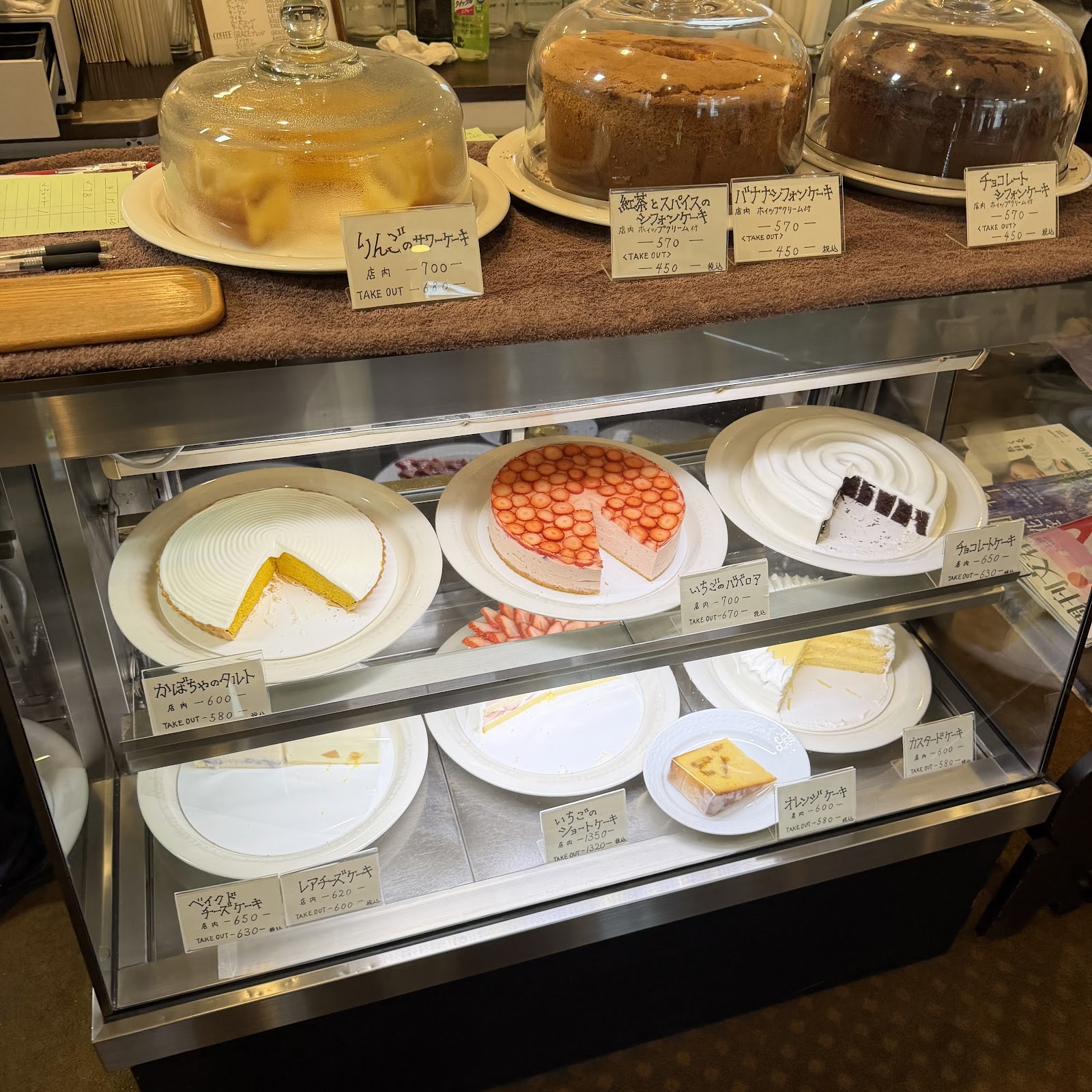 ショーケースに並ぶ季節のケーキ。いちごのババロア、レアチーズ、いちごのショートケーキ、チョコレートなど。値札に手書きの価格と TAKE OUT 表記。