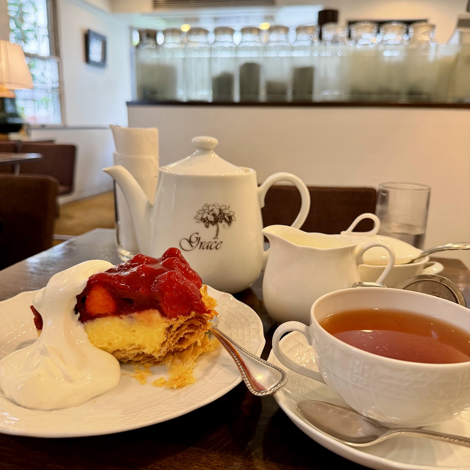 窓辺のテーブルに置かれたいちごのパイと、Grace 印の白い紅茶ポット。背景に紅茶器具棚。